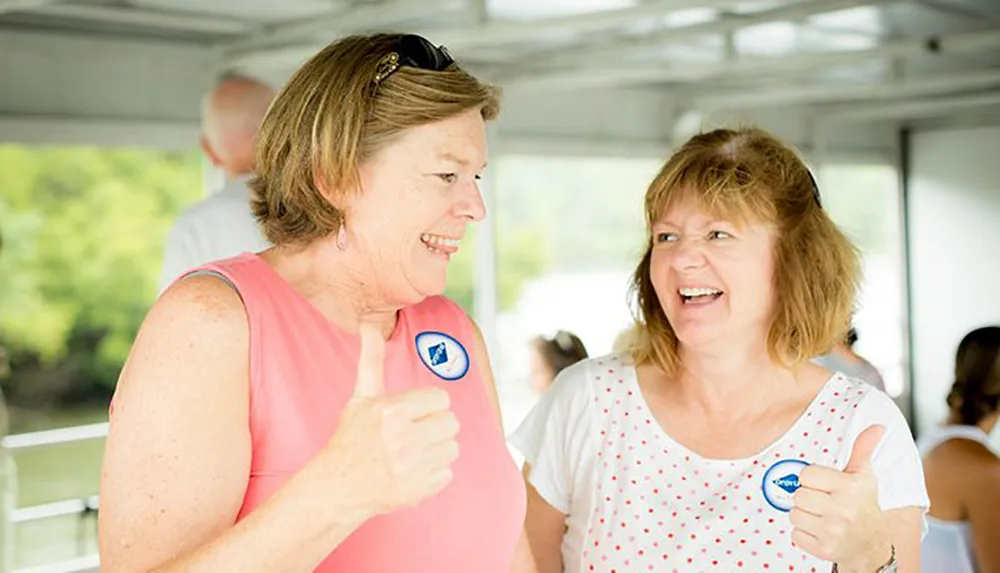 Two women are sharing a joyful moment with thumbs-up gestures wearing stickers displaying a symbol with a person in a circle potentially indicating participation in an event or support for a cause