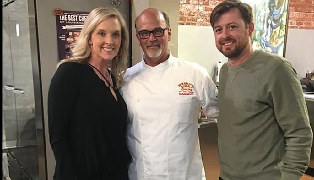 Two individuals are posing for a photo with a chef who is in the center wearing a white chefs coat with embroidery on the left side in what appears to be a restaurant or a cooking environment