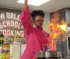 A chef is performing a flamb cooking technique creating a large flame in a pan in a kitchen with a sign reading New Orleans School of Cooking in the background
