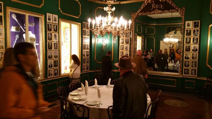 This image depicts a grand dining room with green walls adorned with numerous framed photos, an ornate chandelier, a large mirror, and people milling about, giving the impression of an elegant event or gathering.