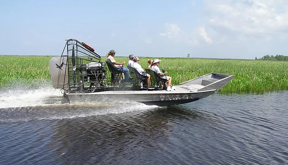 An airboat glides across a waterway surrounded by tall grasses carrying several passengers on what looks like a guided tour