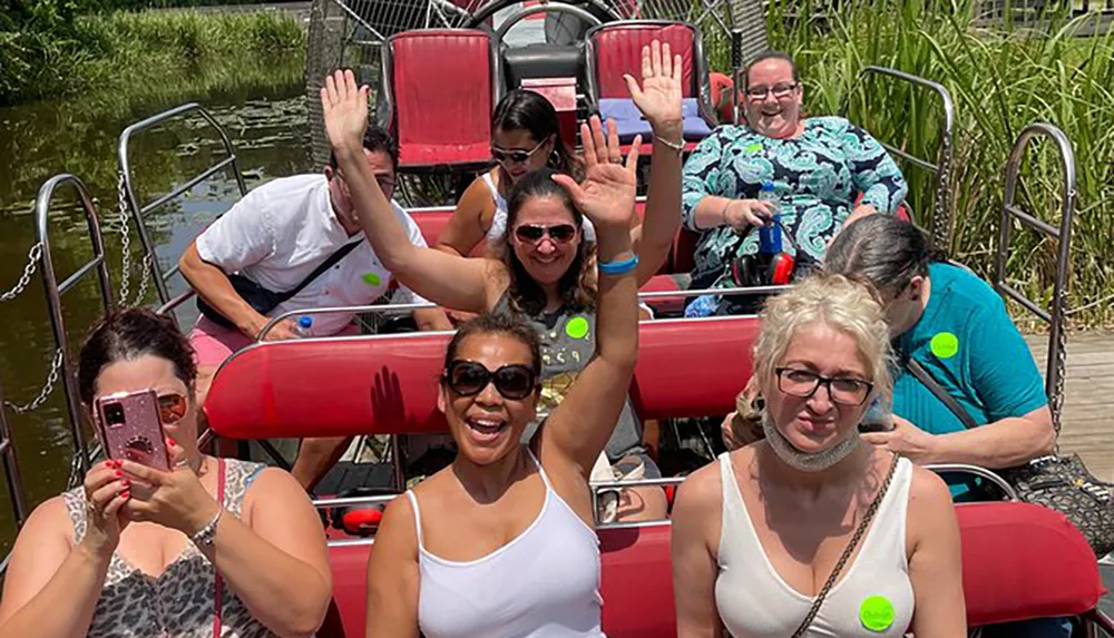A group of cheerful people are sitting in a red boat with some of them waving at the camera capturing a moment of enjoyment on a sunny day