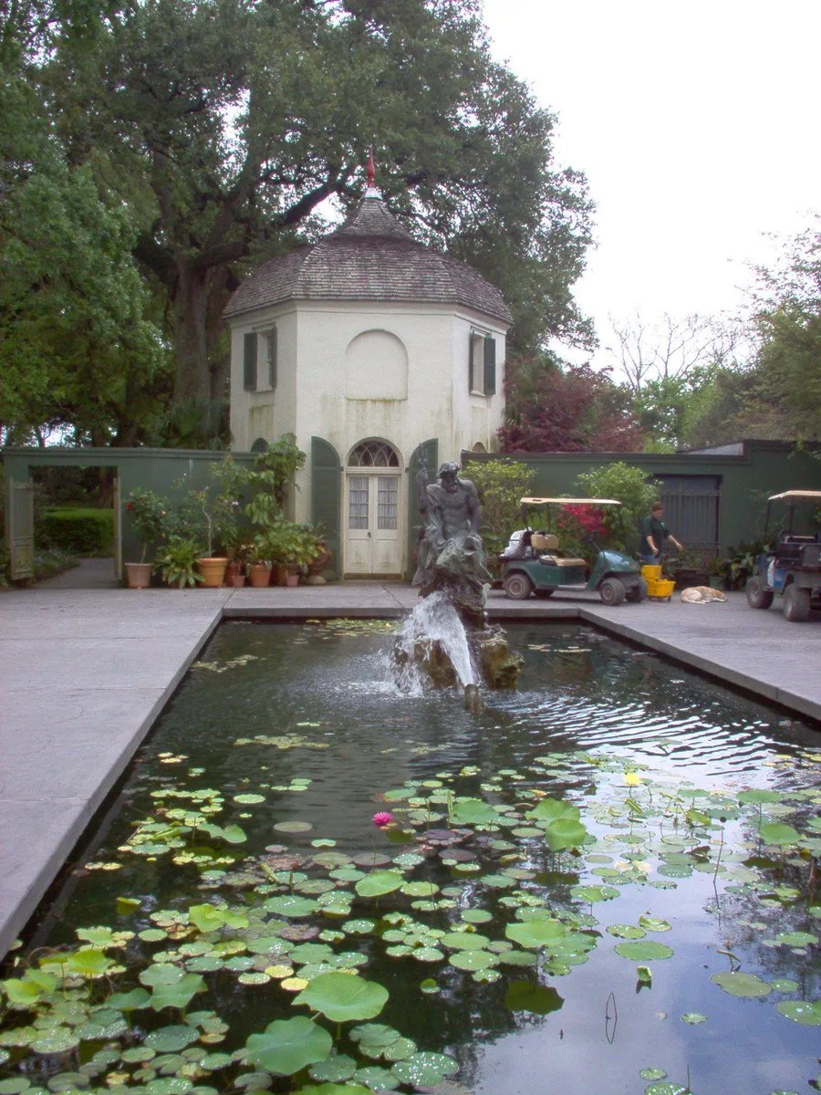 A serene garden scene with a reflective pond featuring water lilies, a fountain statue, and a quaint garden structure in the background.
