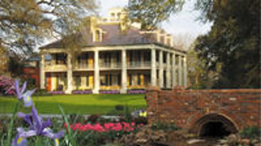 The image shows a stately historical mansion with columns set against a backdrop of mature trees featuring a large lawn in the foreground with colorful flowers and an ornate brick bridge