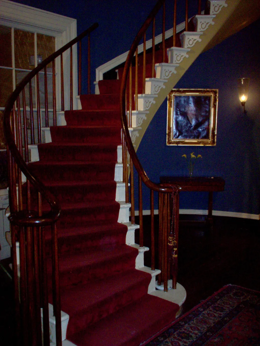 The image shows an elegant staircase with red carpeting ornate white balusters and dark wood handrails complemented by blue walls and a framed picture on the wall