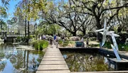 Visitors enjoy a sunny day at a scenic park with a reflective pond, modern sculpture, and Spanish moss-draped trees.