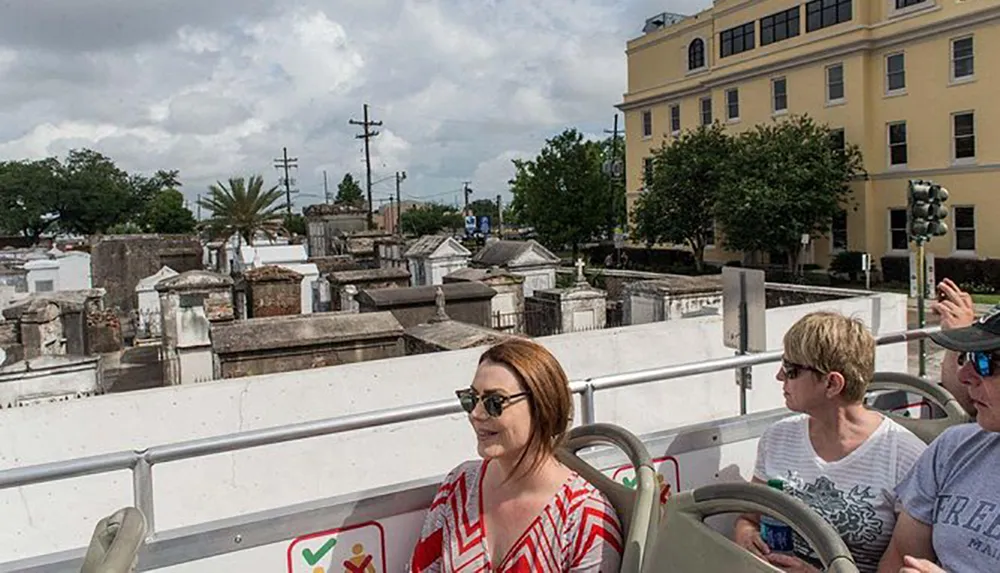 Passengers are seated on an open-top tour bus passing by an old cemetery with above-ground tombs