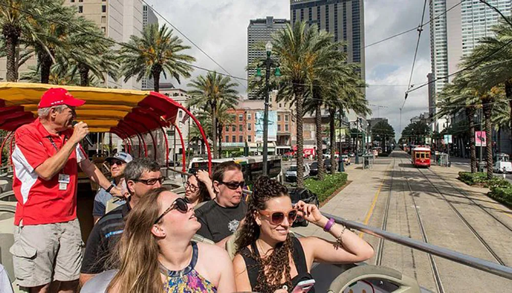 Tourists are enjoying a guided tour on a double-decker bus through a city street lined with palm trees and a streetcar in the background