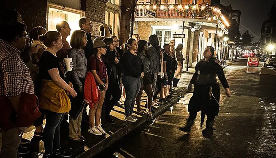 A group of people stands attentively on a sidewalk at night, listening to a guide who seems to be telling a story or giving a tour.