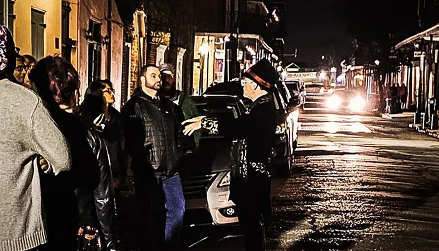 A group of people is engaging with a person in flamboyant attire, possibly a tour guide, on a dimly lit street at night.