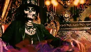 A person with skull face makeup is seated in an ornate chair with purple smoke around, evoking a mystical or Gothic atmosphere.