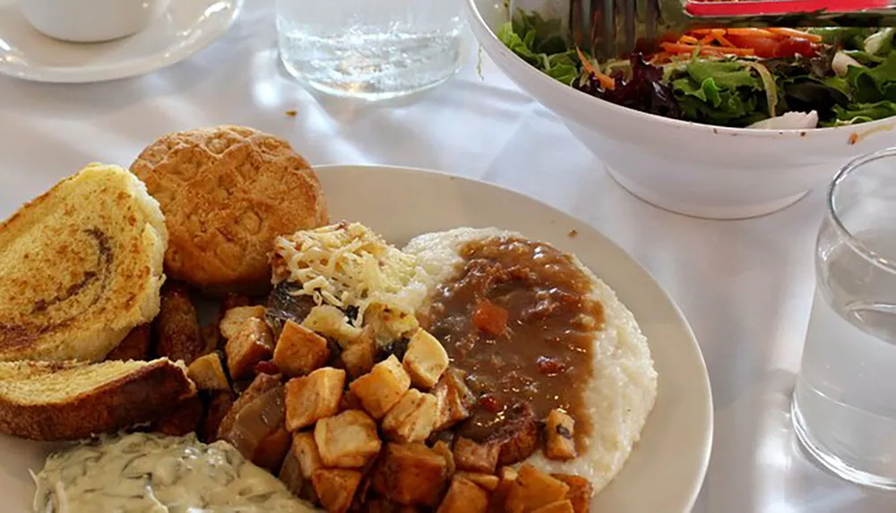 The image shows a hearty meal composed of biscuits gravy grits diced potatoes toast and a side salad arranged on a table set for dining