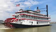 The image depicts the City of New Orleans riverboat, a traditional paddlewheel steamboat, docked on the water under a clear sky with American and state flags flying atop.
