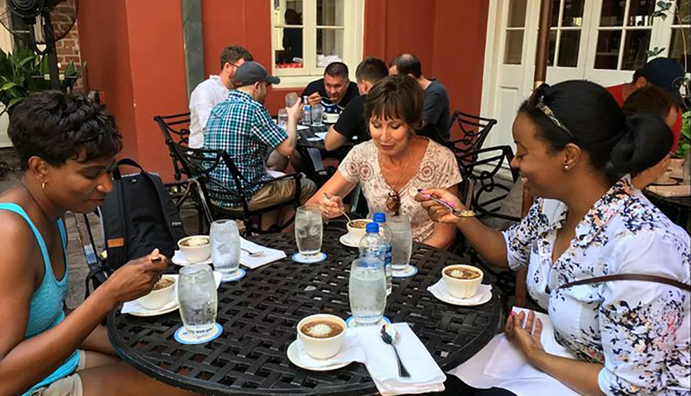 Three people are enjoying a meal outdoors at a cafe with other patrons in the background
