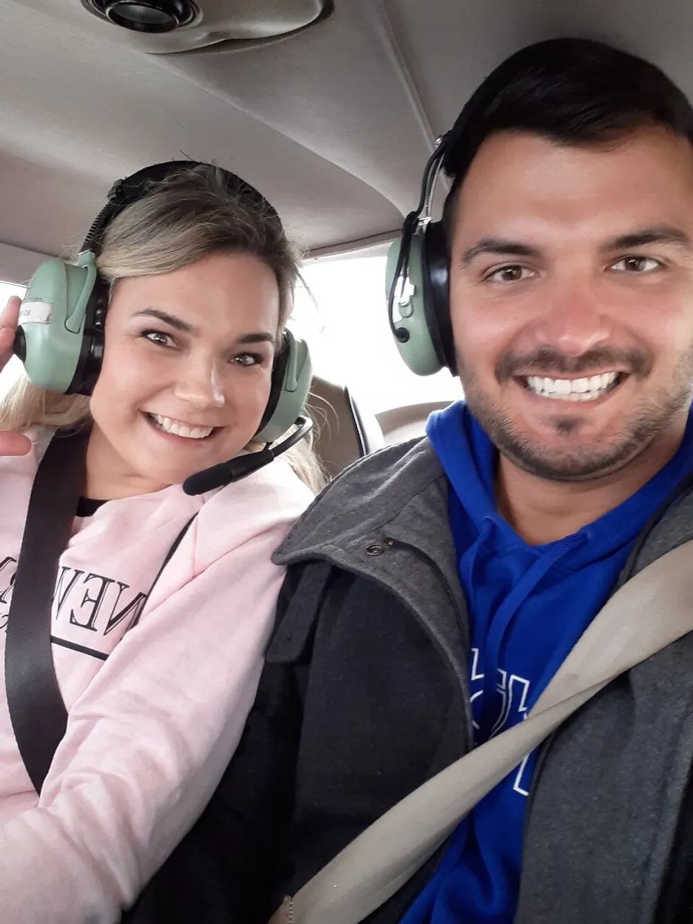 A man and a woman are smiling for a selfie while wearing aviation headsets possibly sitting inside an aircraft