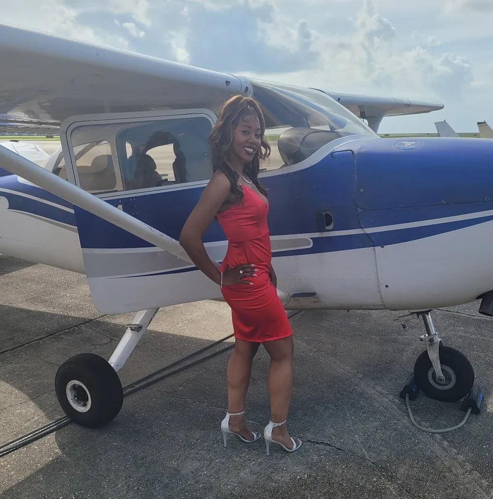 A person in a red dress stands smiling in front of a small plane striking a confident pose