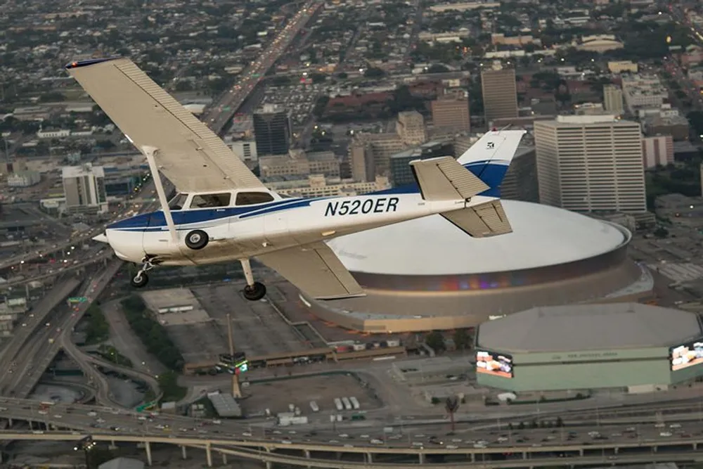 A single-engine light aircraft is flying over an urban area with a large arena in the background