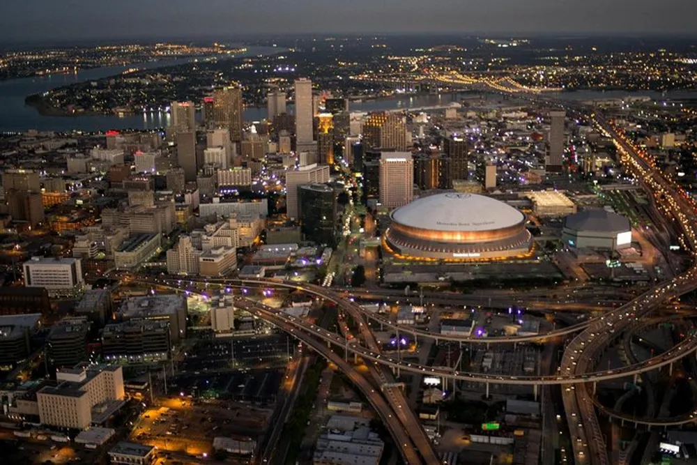 An aerial nighttime view of a bustling city with skyscrapers lit streets and a prominent circular stadium intersected by a network of highways beside a wide river
