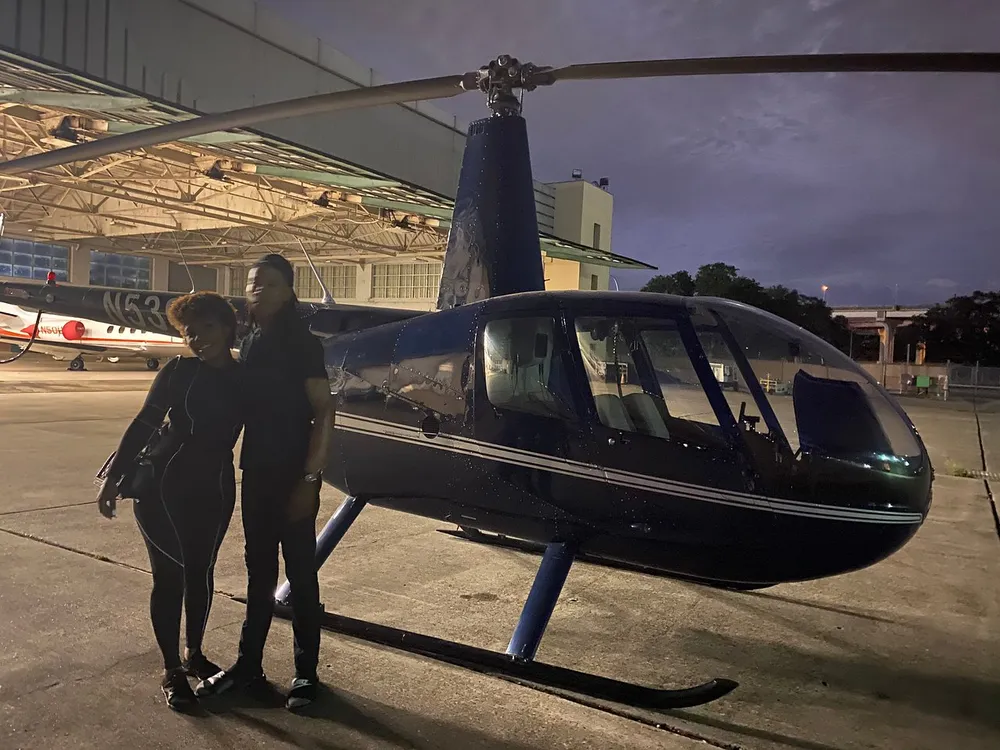 Two people are standing in front of a helicopter at dusk with an airplane and hangar visible in the background