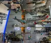 The image shows the interior of an aviation museum with several aircraft suspended from the ceiling and military vehicles on the floor as visitors explore the exhibits
