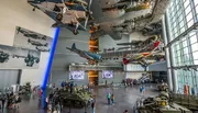 The image shows the interior of an aviation museum with several aircraft suspended from the ceiling and military vehicles on the floor, as visitors explore the exhibits.