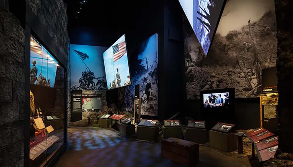 The image displays an interior view of a dimly-lit historical museum with multimedia exhibits including large-scale photographs and video screens telling the story of past military events