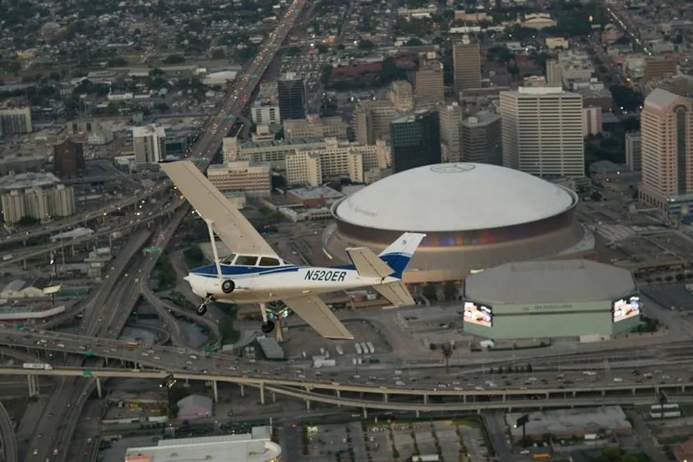 A single-engine aircraft flies over an urban area with a distinctive domed structure and intersecting roadways