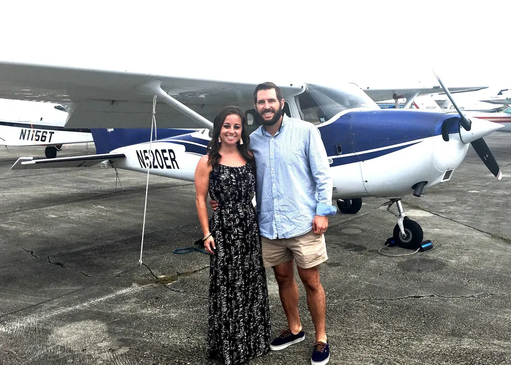 A smiling woman and man are standing in front of a small airplane parked on a tarmac
