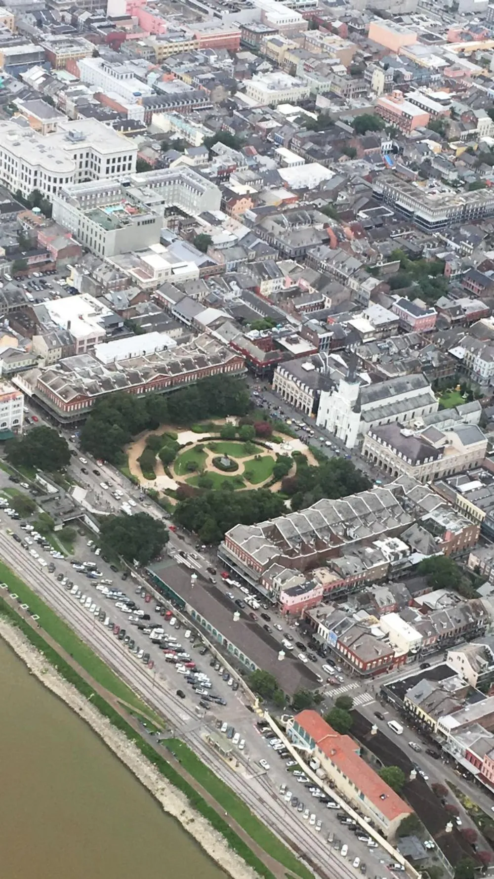 The image is an aerial view of a densely built urban area with distinctive architecture featuring a large symmetrical green space surrounded by historic buildings adjacent to a highway and a body of water