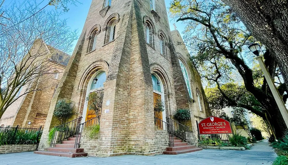 This is an image of the St Georges Episcopal Church featuring its brick architecture and a sign welcoming visitors near a lamp post with trees and a clear sky in the background