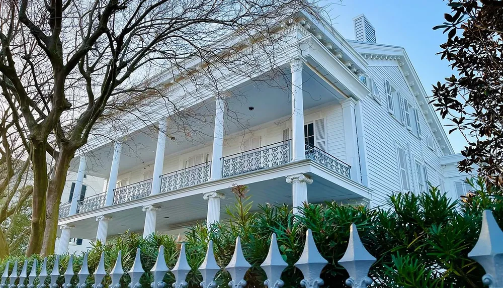 The image shows a grand white house with double-tiered balconies featuring ornate railings framed by leafless tree branches and a decorative iron fence