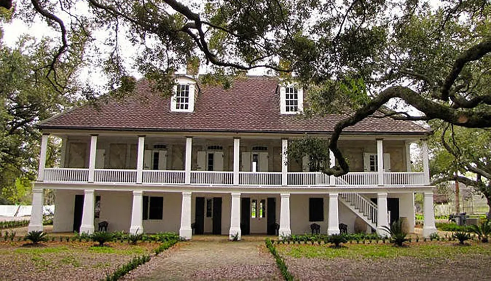 The image shows a two-story white plantation-style house with a covered porch on each level set among mature oak trees draped with Spanish moss