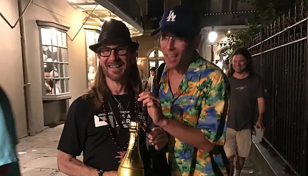 Two individuals are happily posing with a trophy on a lively street at night one wearing a vibrant tropical shirt and a baseball cap and the other in a black T-shirt and a fedora with a third person smiling in the background