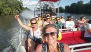 Passengers are enjoying a sunny airboat tour, with several wearing ear protection, possibly indicating the high noise levels of the boat engine.