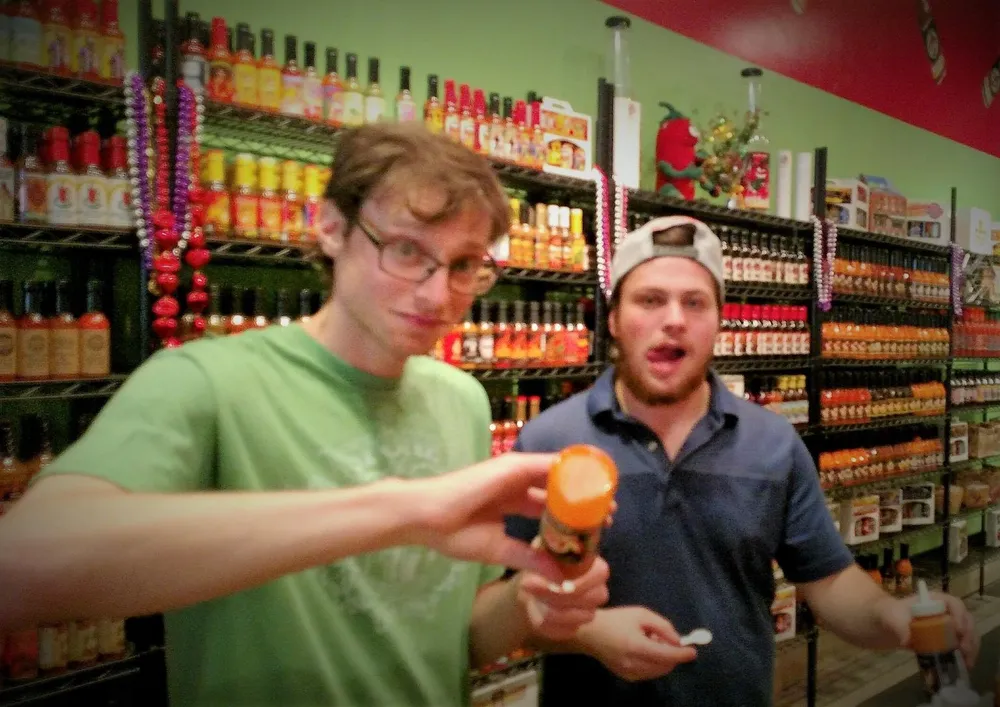 Two people stand in front of shelves packed with various bottles of hot sauce with one holding a hot sauce bottle and the other person looking surprised or excited