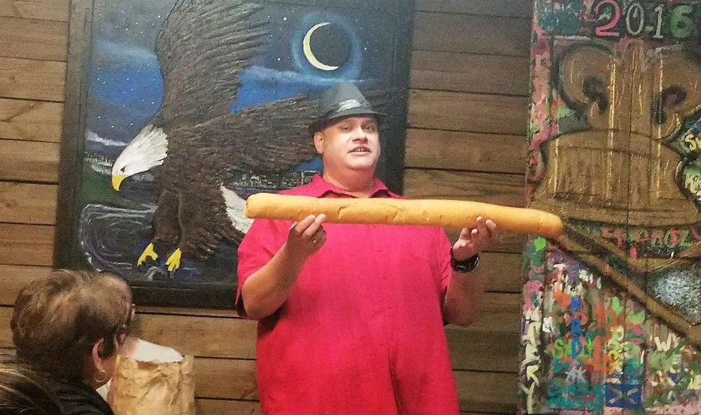 A man wearing a black hat and a red shirt is holding a large breadstick with a painting of an eagle in the background