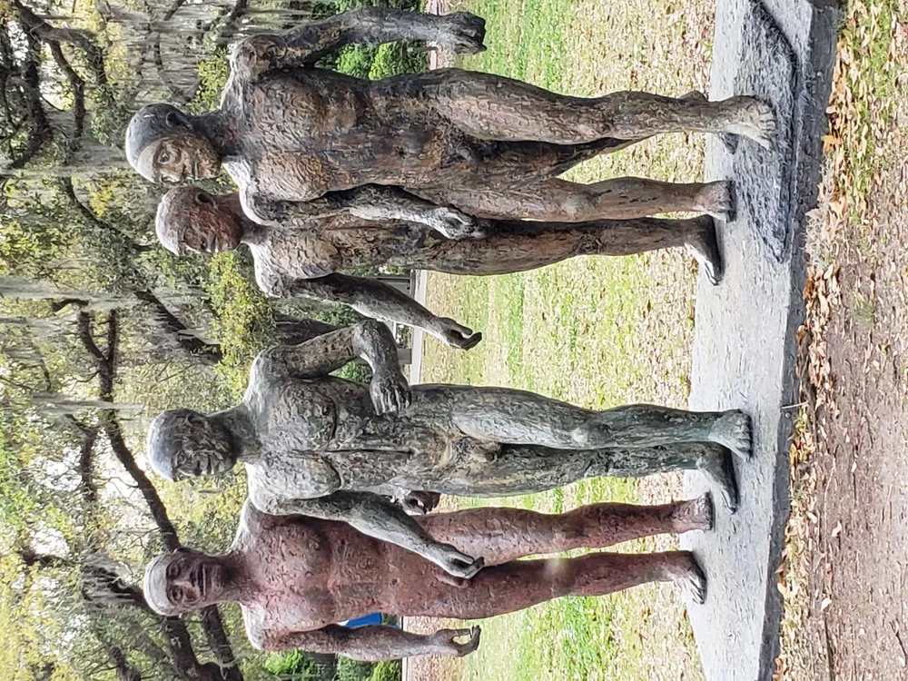 The image shows a group of weathered bronze statues of people with expressive poses standing on a stone platform in an outdoor setting surrounded by trees