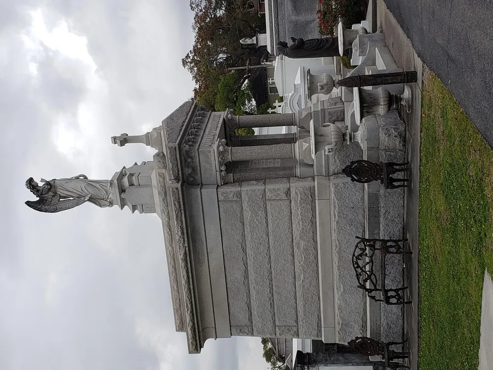 The image shows a majestic angel statue as part of a larger grave monument under an overcast sky but the photo is rotated 90 degrees to the right