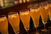 A row of champagne flutes filled with a bubbling golden-colored beverage, possibly champagne or sparkling wine, is arranged on a bar counter with a softly lit background suggesting a warm ambiance.