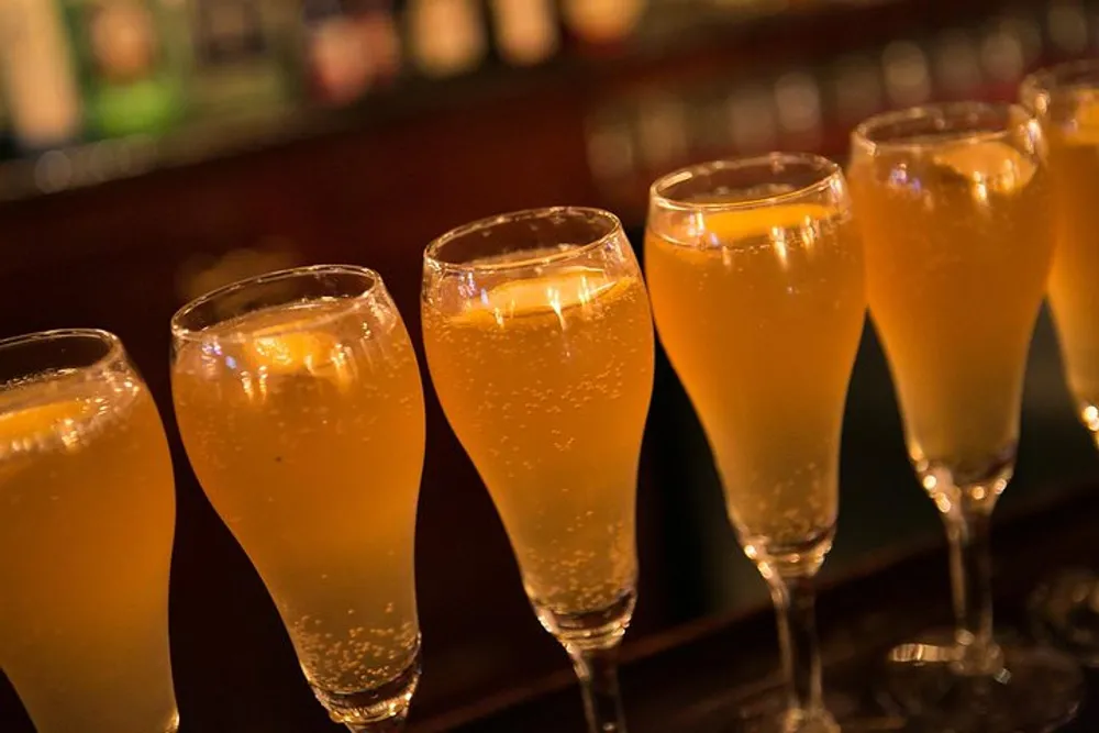 A row of champagne flutes filled with a bubbling golden-colored beverage possibly champagne or sparkling wine is arranged on a bar counter with a softly lit background suggesting a warm ambiance