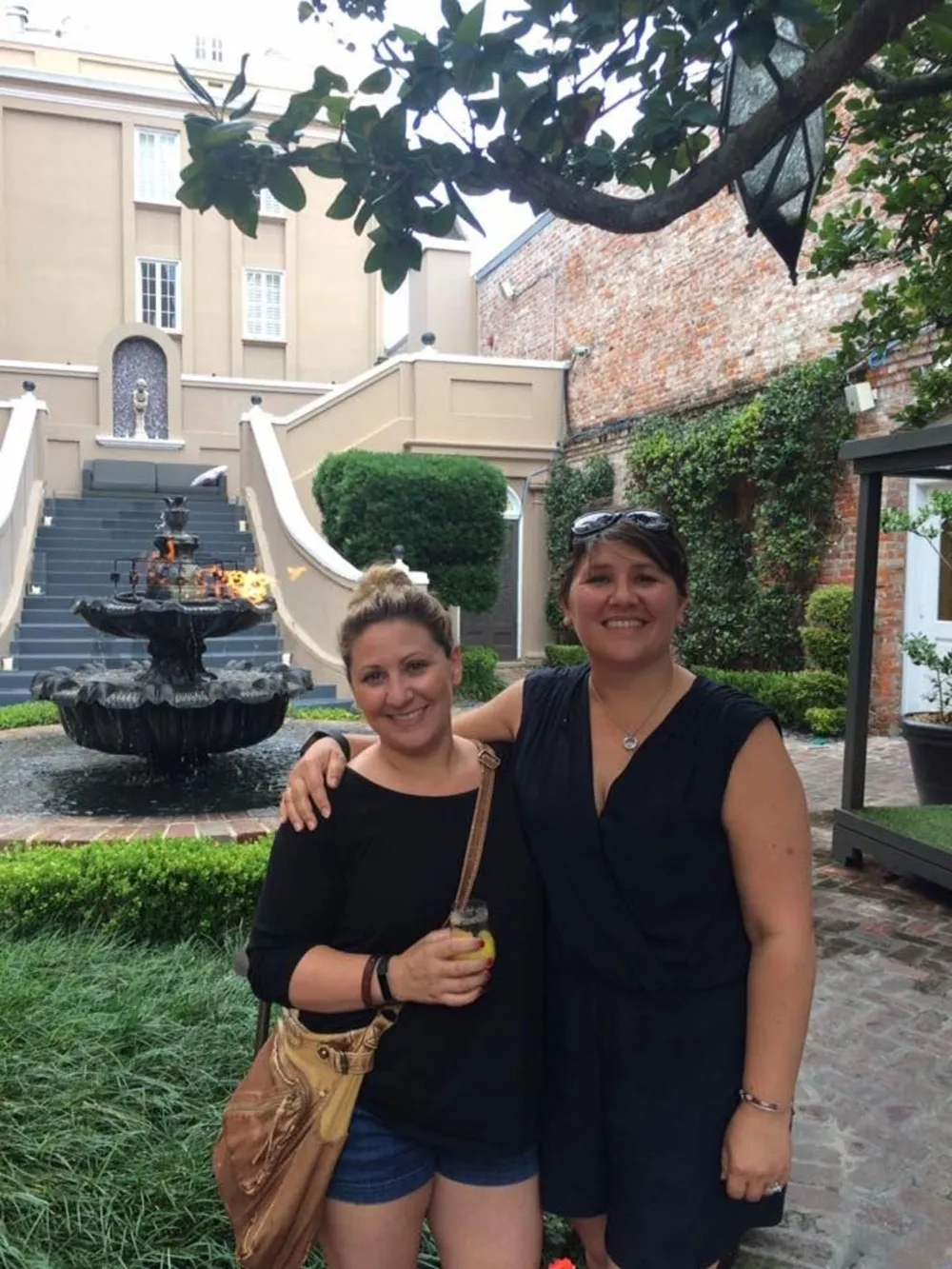 Two smiling women are posing in front of an ornate fountain in a charming courtyard setting with greenery and a classic architectural backdrop