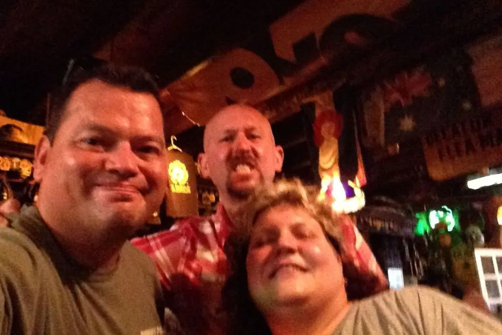 Three people are taking a friendly selfie together in a pub-like setting with warm lighting and eclectic decorations in the background