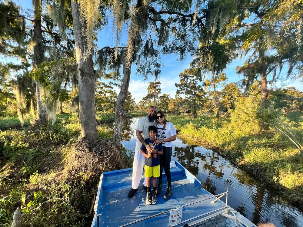 Three people are standing together smiling on a boat surrounded by a serene waterway with lush greenery and hanging Spanish moss