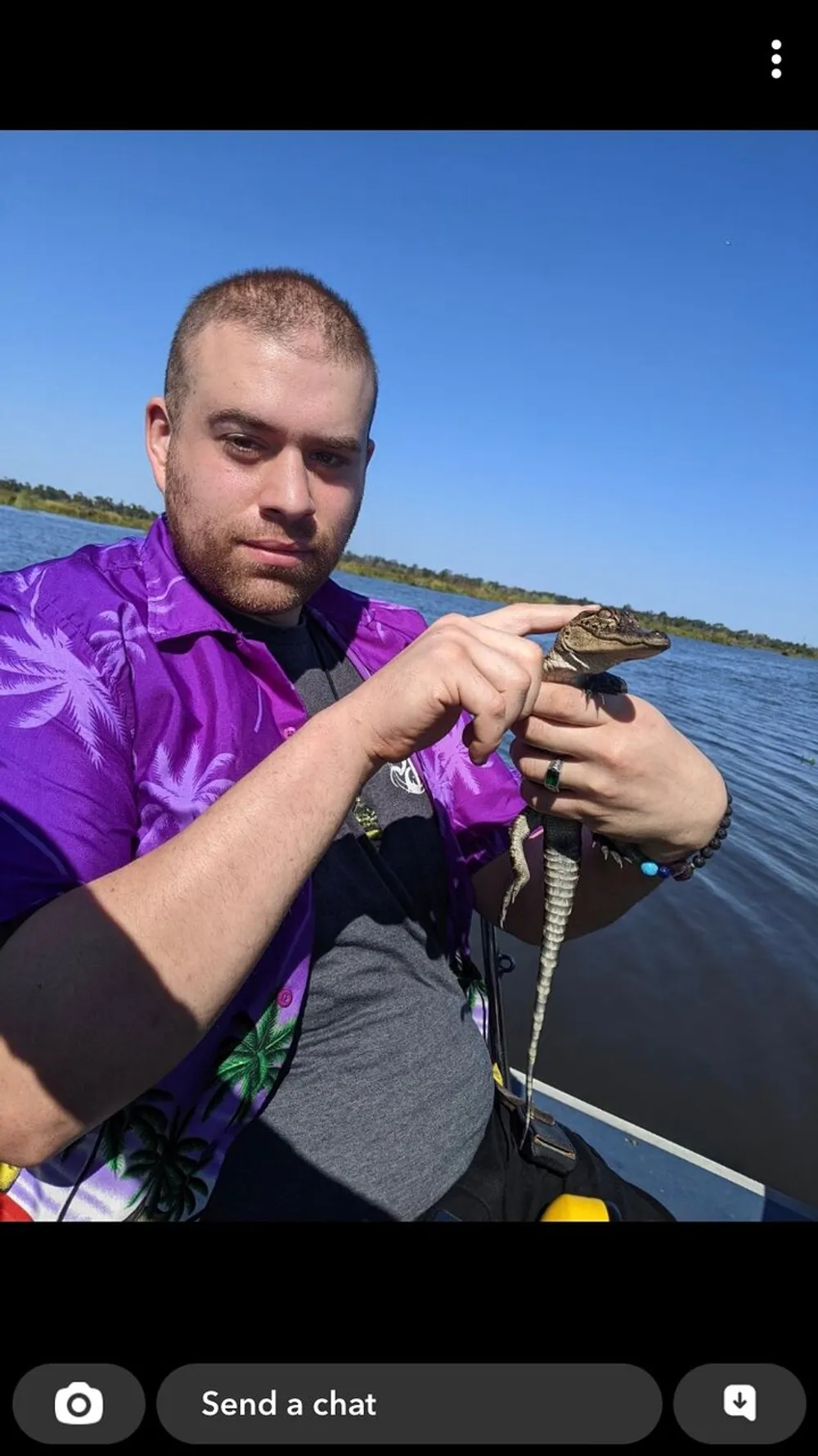 A person in a purple shirt is holding a small alligator on a boat with a lake and clear sky in the background