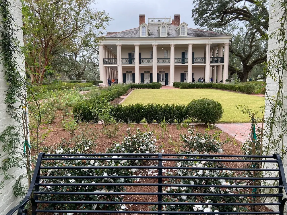 This image shows a stately two-story house with a large porch and columns overlooking a manicured garden with a lawn and flowering bushes as seen over a metal bench
