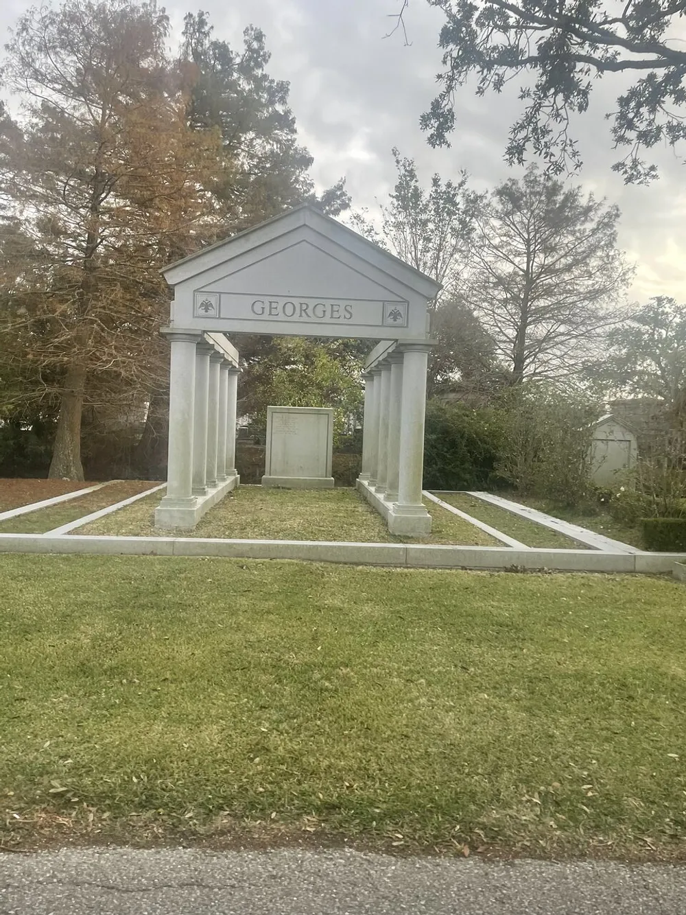 The image shows an elegant classical-style family mausoleum with the name GEORGES inscribed at the top surrounded by trees under a cloudy sky