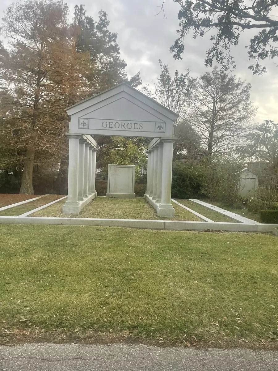 The image shows an elegant, classical-style family mausoleum with the name GEORGES inscribed at the top, surrounded by trees under a cloudy sky.