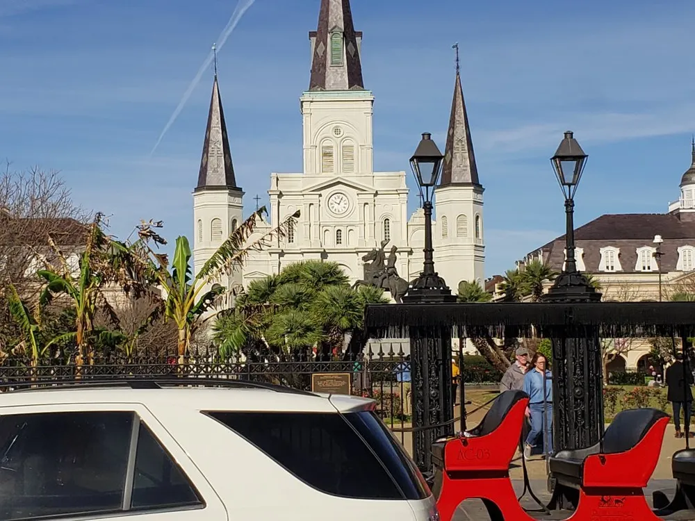 The image shows a view of the historic St Louis Cathedral seen from Jackson Square with a glimpse of passing pedestrians street lamps tropical plants and the front part of parked vehicles in the foreground