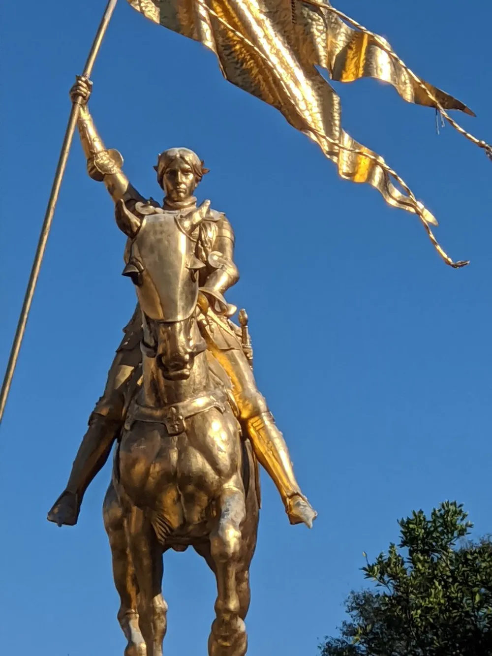 The image shows a golden statue of a mounted figure in armor holding aloft a flag that flutters in the breeze against a clear blue sky