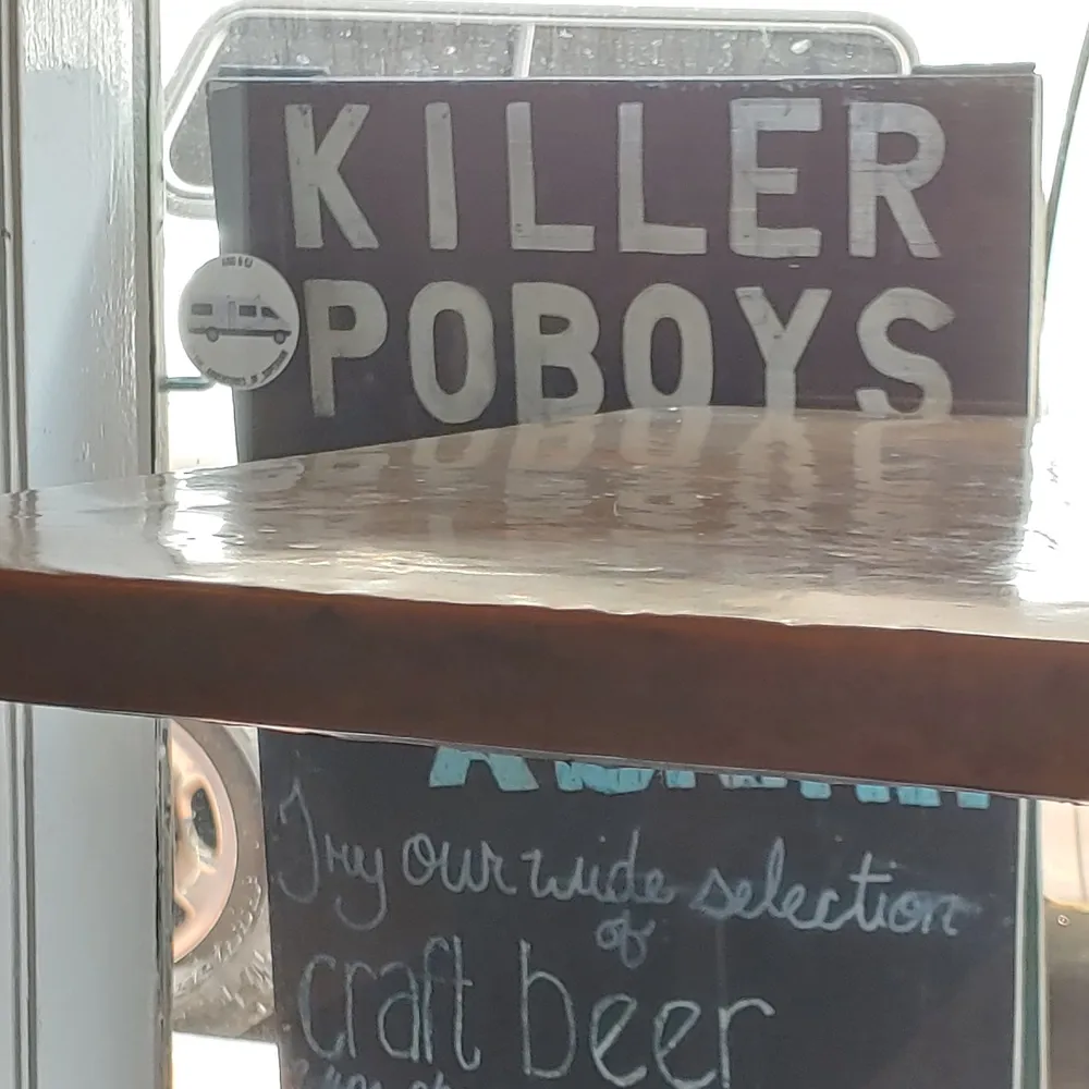 The image displays a reflective table surface with a sign in the background that reads KILLER POBOYS and part of another sign visible at the bottom that suggests a selection of craft beer